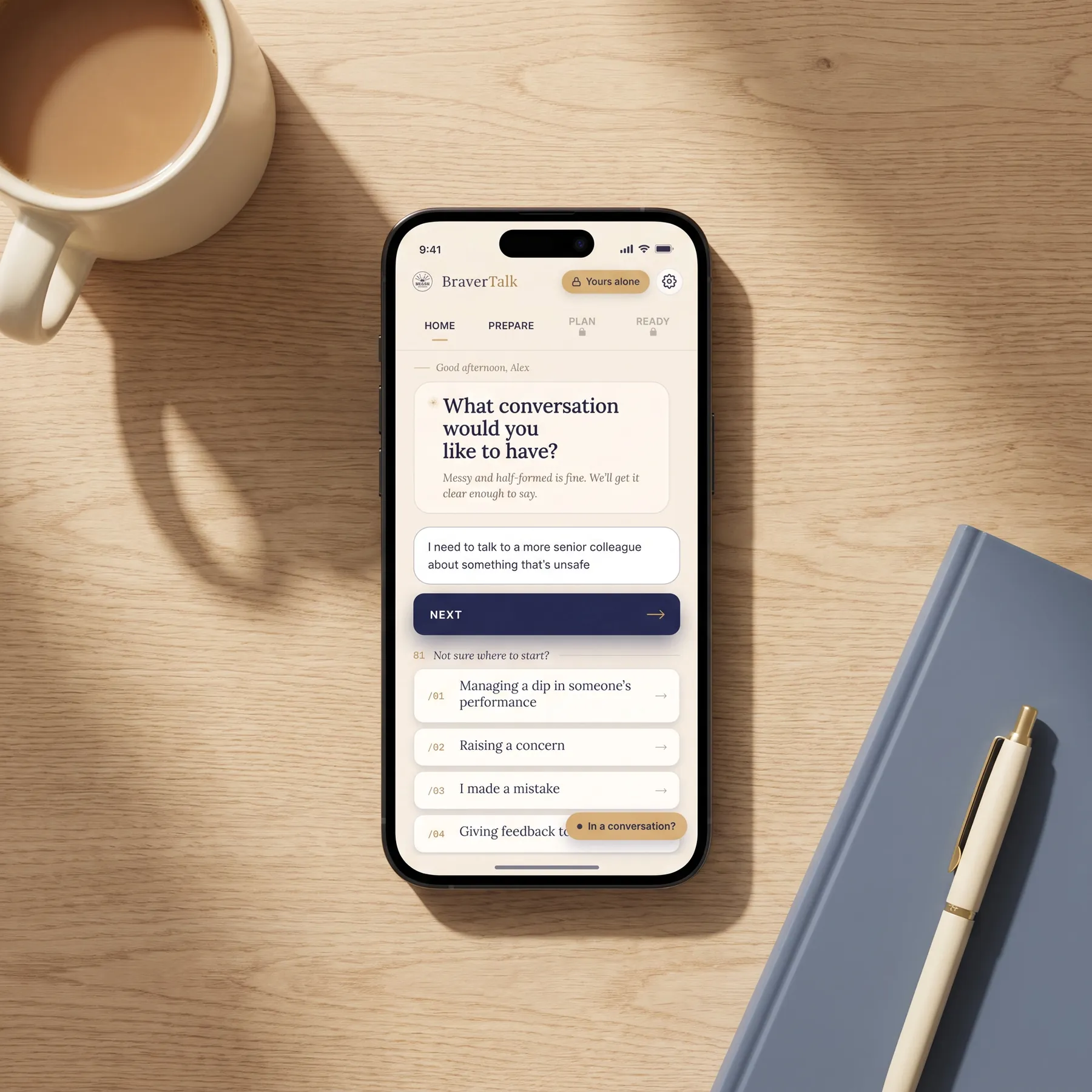 The BraverTalk app open on a phone on a wooden desk, asking &lsquo;What conversation would you like to have?&rsquo;, beside a mug of tea and a notebook